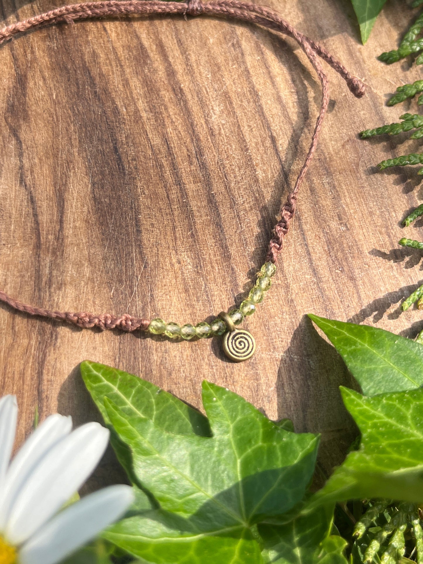 anklet with a spiral charm on a wooden surface surrounded by green leaves and a white flower.