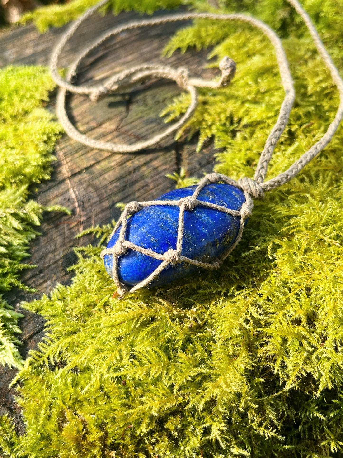 Handmade macrame necklace with an Lapis Lazuli crystal wrapped in hemp cord, featuring gemstone beads and spiral metal accents. Bohemian jewellery displayed on green moss with a rustic wood background