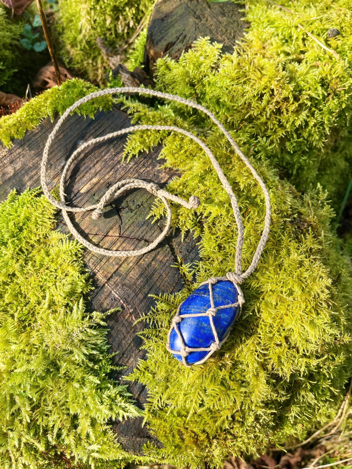 Handmade macrame necklace with an Lapis Lazuli crystal wrapped in hemp cord, featuring gemstone beads and spiral metal accents. Bohemian jewellery displayed on green moss with a rustic wood background