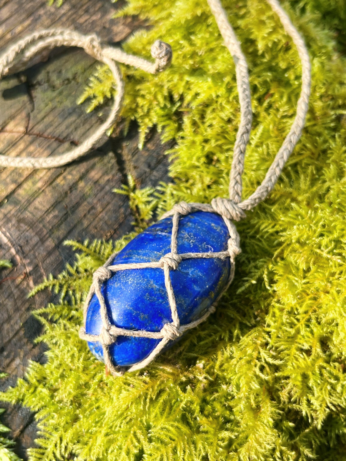 Handmade macrame necklace with an Lapis Lazuli crystal wrapped in hemp cord, featuring gemstone beads and spiral metal accents. Bohemian jewellery displayed on green moss with a rustic wood background